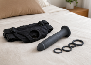 A slim harness-compatible silicone dildo laid beside interchangeable O-rings and a folded cotton harness on a bed.