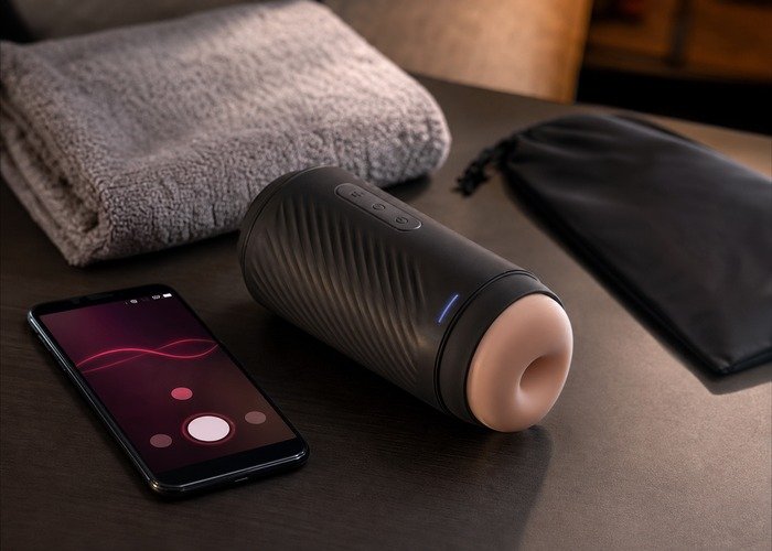 A realistic automatic male masturbator, smartphone, charger, towel, and pouch arranged on a bedside table for an app-control guide.