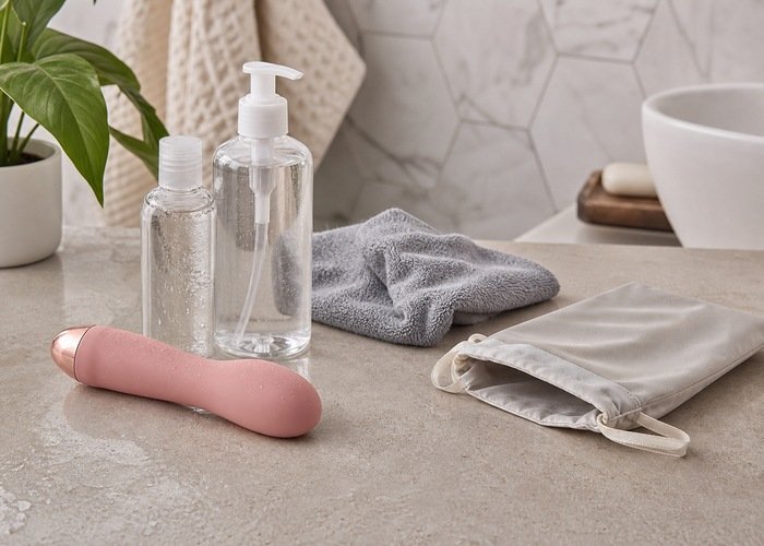 A silicone adult wellness vibrator beside unbranded lubricant bottles, a towel, and a storage pouch on a bathroom counter.
