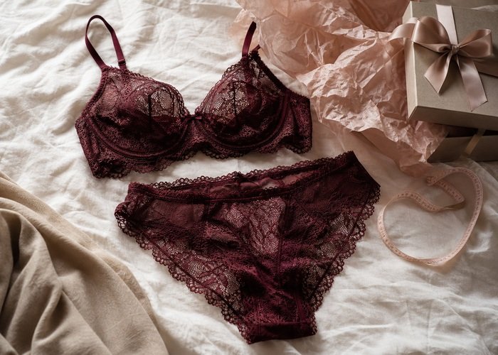 A realistic lace lingerie set, gift box, soft measuring tape, and tissue paper arranged on a bed for a gift guide.