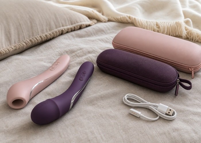 Two oral-style adult wellness vibrators with distinct rounded contact heads arranged with pouches on a linen bed.