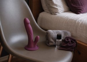 A realistic suction-cup dildo, lubricant, folded towel, and storage pouch arranged on a bed for a surface stability guide.