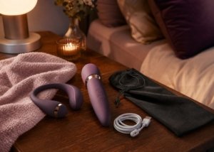 Two discreet adult wellness products arranged with a towel, charging cable, and storage pouch on a bedside table.