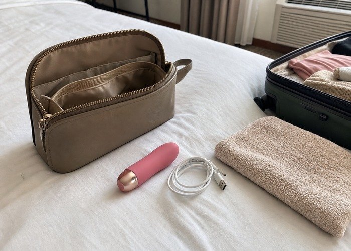A close bedside scene with a compact vibrator, pouch, charging cable, and folded towel arranged to explain packing and lock checks.