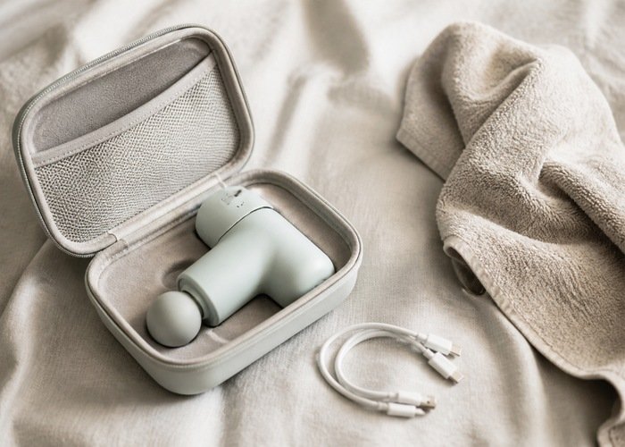 A personal wellness device in an open travel case beside a towel and cable on a linen bed.