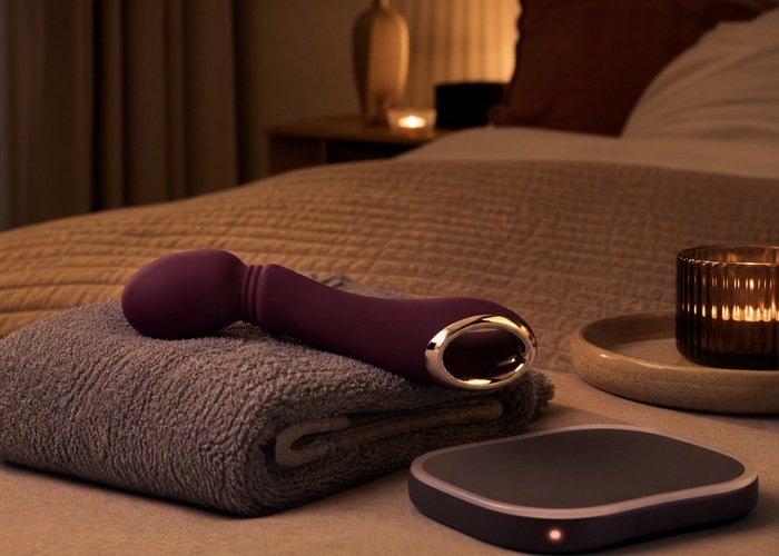 A silicone adult wellness vibrator beside a towel and simple warming accessory in a softly lit bedroom.