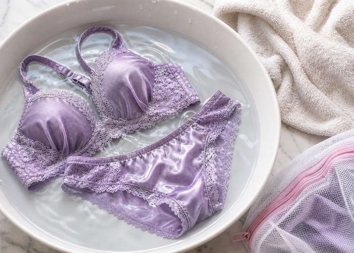 A satin and lace lingerie set soaking in a shallow basin with cool water, soft towel, and mesh bag nearby.