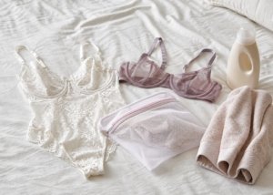 A realistic lace bodysuit, mesh bra, and laundry bag arranged beside delicate detergent and a folded towel.