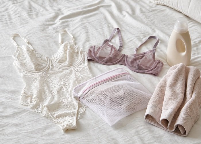 A realistic lace bodysuit, mesh bra, and laundry bag arranged beside delicate detergent and a folded towel.